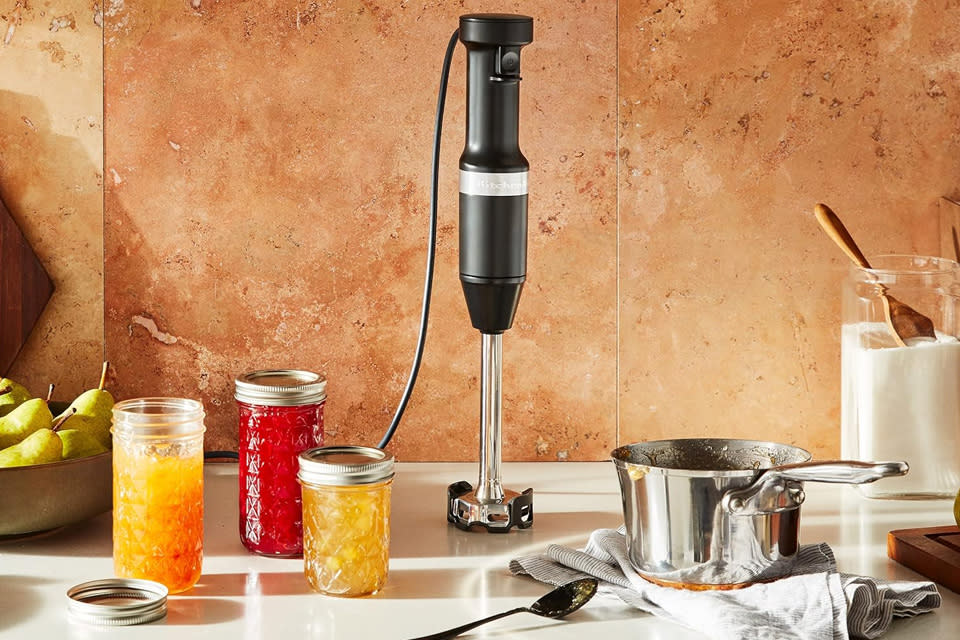 KitchenAid Variable Speed Corded Hand Blender stands on a coutertop with various blended foods nearby