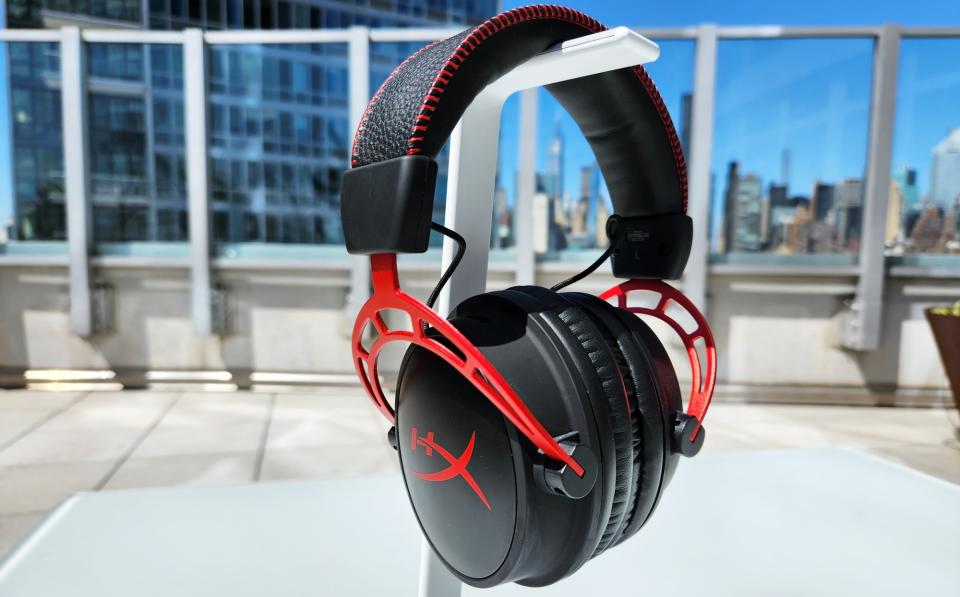 A black and red gaming headset, the HyperX Cloud Alpha Wireless, rests on a white headphone stand on a table outdoors.