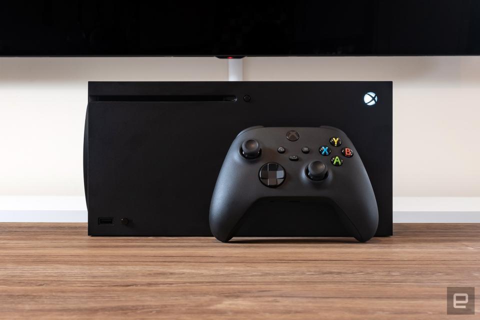 The Xbox Series X game console lays on its side, with a black Xbox controller rested in front of it, on top of a brown wooden desk.